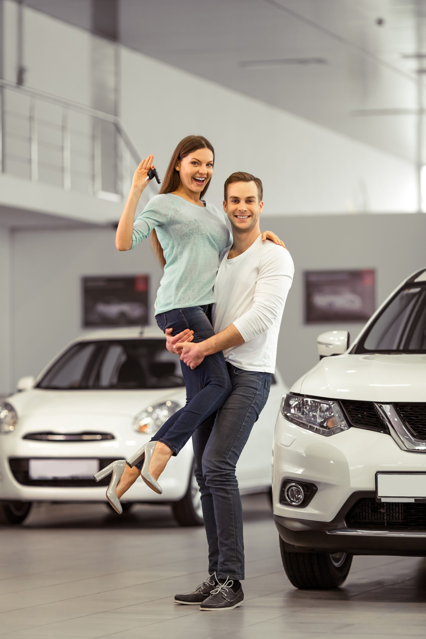 Beautiful young couple is smiling, looking at camera and showing keys while buying a car in a motor show. Man is holding woman in his arms